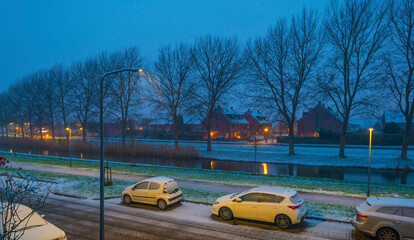 First snow at night in a residential street with parked cars along a canal in winter, Almere,...