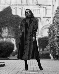 Stylish woman in red long coat with belt, black skirt, tights, sunglasses and high heels walking at city street. Brunette caucasian girl with long wavy hair outdoor portrait