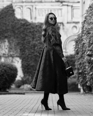 Stylish woman in red long coat with belt, black skirt, tights, sunglasses and high heels walking at city street. Brunette caucasian girl with long wavy hair outdoor portrait