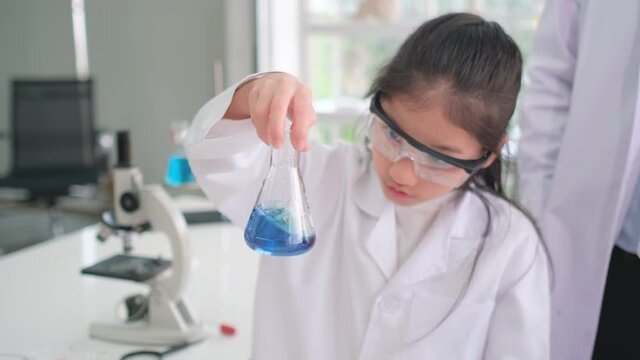 Young scientist girl express exciting after experiment with blue chemical in flask in laboratory or classroom with teacher and classmate.