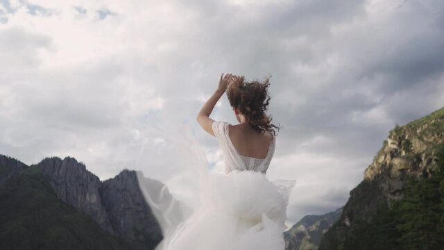 A bride in a wedding dress flying in the wind stands on a rocky river bank against the backdrop of mountains in Altai