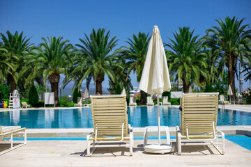 Two white plastic sun loungers and parasol stand near the pool with turquoise water on the pallm trees background