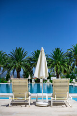Two white plastic sun loungers and parasol stand near the pool with turquoise water on the pallm trees background, vertical orientation