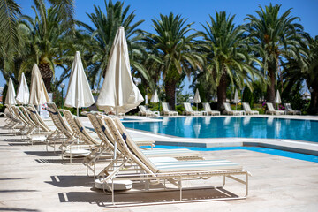 Row of white plastic sun loungers and parasols near the pool with turquoise water on the palm trees background