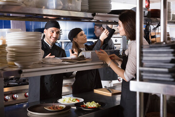 Happy positive smiling woman waiter is giving order to chef cook on kitchen in restaurant.