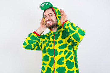 Young caucasian man wearing a pajama standing against white background Trying to hear both hands on ear gesture, curious for gossip. Hearing problem, deaf