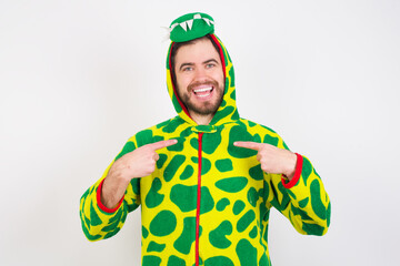 Fototapeta premium Young caucasian man wearing a pajama standing against white background points at his body, being in good mood after going shopping and making successful purchases