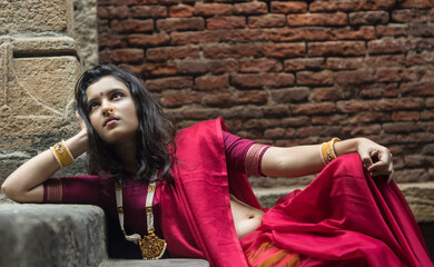 Portrait of beautiful Indian girl in heritage stepwell wearing traditional Indian red saree, gold...