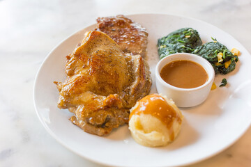 Pork collar steak and chicken steak served with corn spinach, mashed potatoes and brown sauce which put on a white plate on marble table..