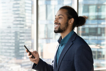 Happy millennial African American businessman use cellphone look in distance dreaming or thinking in office. Smiling biracial male CEO or employee hold browse modern smartphone, plan imagine success.