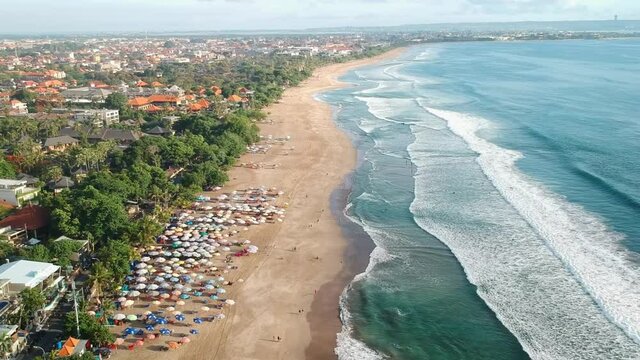 Aerial flyover sandy Legian Beach with beautiful landscape during sunlight, waves crashing on shore and many sunshades. Bali,Indonesia.