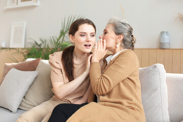 Naklejka na ściany i meble Happy adult daughter whispering secret to her middle mother at home, gossiping, sharing secret. Beautiful woman hearing good news from mature mum, trusted relations.
