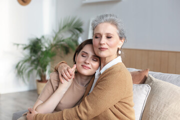 Cheerful young woman is embracing her middle aged mother with closed eyes hugging, touching cheeks, sitting on couch at home. Happy trusted relations. Family concept.