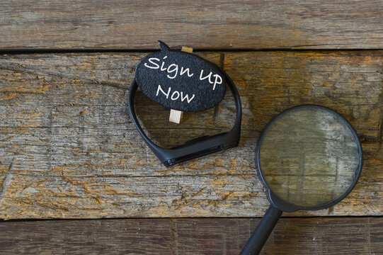 Top view of magnifying glass, watch and wooden tag written with text SIGN UP NOW over wooden background. Business concept.