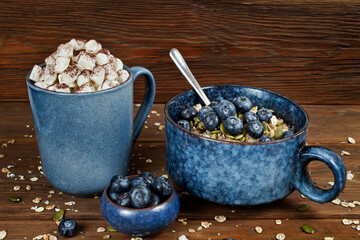 Oatmeal porridge with chia and pumkin seeds, blueberries  in a blue ceramic bowl and cup of cocoa drink with marshmallows on a wooden background. Healthy eating and tasty breakfast concept.