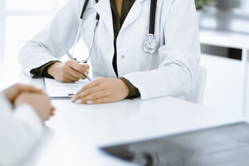 Unknown woman-doctor and female patient sitting and talking at medical examination in clinic, close-up. Therapist wearing green blouse is filling up medication history record. Medicine concept