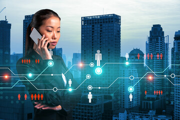 Attractive Asian businesswoman processing conference call to develop social media marketing strategy to achieve business goals. Hologram icons over Bangkok background.