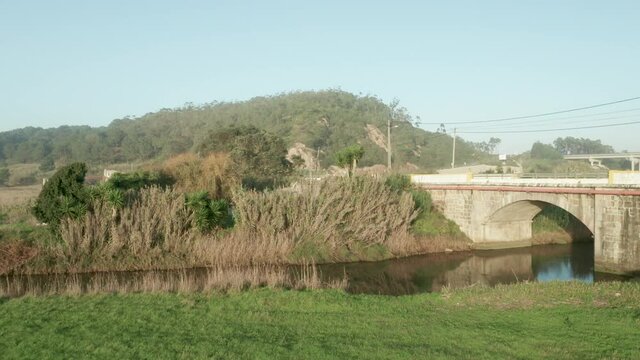 Car Traveling Across Stone Arch Bridge Over Calm Lake Of Alcobaca River Near Nazare In Portugal. - Aerial Arc Shot