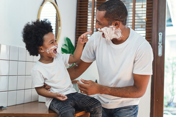 African American father and little son spend time together have a fun laughing while shaving foam on their faces in the bathroom. Morning routine in bathroom concept. - Powered by Adobe