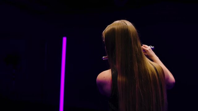 Camera rotates around charming female flutist in a black dress against a background of bright neon lights in a dark studio. Young woman playing melody on a flute. Close up. Slow motion.