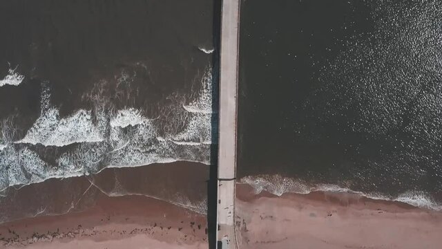 Aerial birds eye view drone footage of a waves crashing onto the beach against a pier, Roker Beach, Sunderland, England.