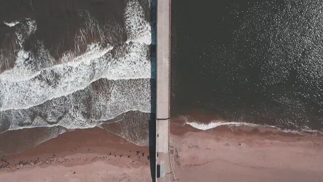 Aerial birds eye view drone footage of a waves crashing onto the beach against a pier, Roker Beach, Sunderland, UK.