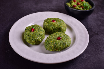 Green pkhali with pomegranate seeds in a white plate on a dark stone table.