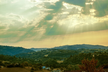 rural mountain landscape with scattered houses, shallow depth of field, mountains and bright sky with sun rays through their clouds.