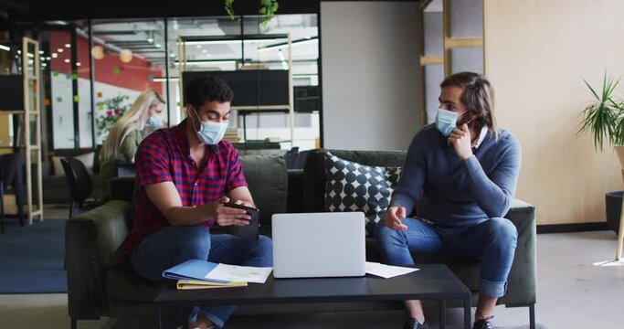 Diverse business people wearing masks using laptop and goign through paperwork in modern office
