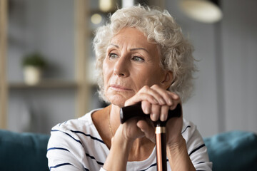 Head shot thoughtful elderly mature grandmother holding hands on walking wooden cane, looking in distance. Unhappy pensive disabled middle aged woman feeling lonely at home, recollecting memories.