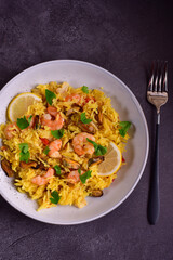 Paella with shrimps, mussels, lemon, herbs in a gray plate with fork on a dark stone table .
