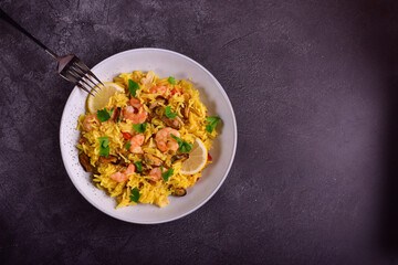 Paella with shrimps, mussels, lemon, herbs in a gray plate next to a fork on a dark gray stone background.