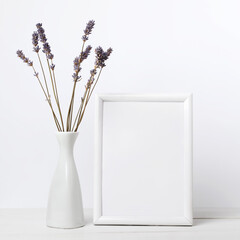 Dried lavender flowers in white vase and white wooden frame with empty paper sheet on wooden surface over light background. Interior floral composition.