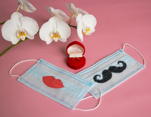 Engagement ring next to disposable protective masks with mustache and lips painted on them on pink background. The concept of St. Valentine's Day, wedding or other holidays during pandemic of COVID-19