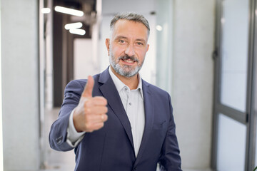 Cheerful handsome middle aged bearded Caucasian businessman, wearing shirt and jacket, showing thumbs up, posing and smiling at camera, in front of modern light loft office corridor