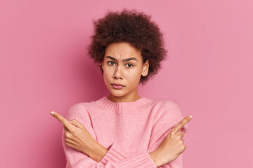 Serious African American woman with curly hair crosses hands and points at different sides hesitates between two items wears casual sweater isolated over pink background. Hard decision concept