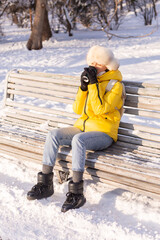 Happy young woman in winter in warm clothes in a snowy park on a sunny day sits on the benches and enjoys the fresh air and coffee alone