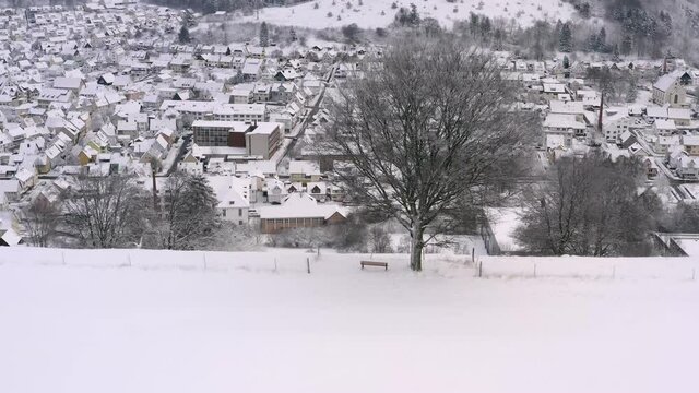 Dronie shot, flaying backwards from a snow covered tree with a bench back to dicover a valley with a snow covered, white and dreamy town.