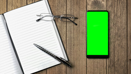 Top view of open notebook with blank pages and cell phone with green screen, glasses on wooden...