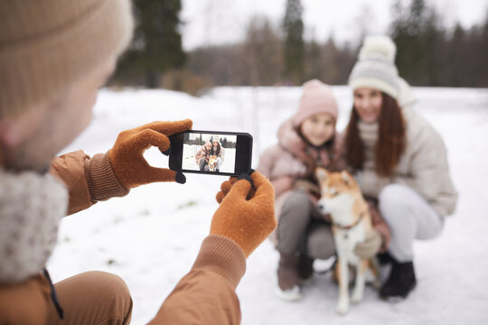 Portrait of father taking photo of cute daughter and wife with dog while enjoying walk outdoors together in winter forest, focus on smartphone screen, copy space