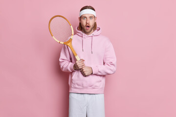 Shocked tennis player holds racket wears headband sweatshirt stunned to lose competition leads active lifestyle poses against pink background enjoys favorite game. Sport and recreation concept