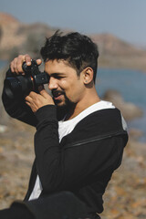 Young indian photographer clicking photos of people near a lake scenery, weraing black hoodie. Enjoyment concept