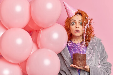 Photo of speechless emotive redhead woman wears birthday hat surprised to see all guests came home holds chocolate cake with bunch of inflated balloons isolated over pink background. Holiday event