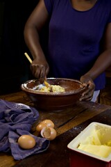 a black person stirring cake batter in a wooden bowl at a kitchen table.  A blue cloth, eggshells and butter sits in the foreground