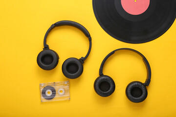 Two pairs of black stereo headphones, vinyl records and audio cassette on yellow background. Top view