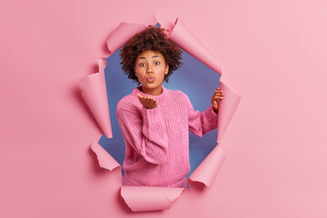Affectionate dark skinned woman blows mwah at camera keeps palm near folded lips expresses love wears knitted sweater breaks through paper background. Tender lovely Afro American girl sends air kiss
