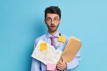 Shocked male student has papers with written math solution greatly surprised to have deadline wears round spectacles formal shirt and tie isolated over blue background. Man works on business plan