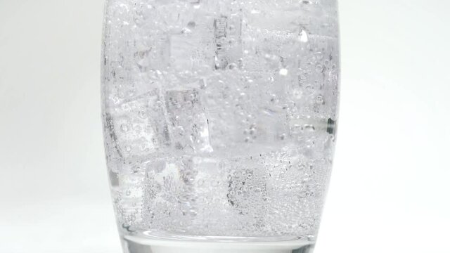 Soda or sparkling water pouring onto ice cubes filling up glass in slow motion bubbles close up carbonation white studio background