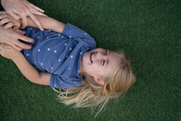 Niña rubia con ojos azules tumbada en el cesped feliz porque unas manos le hacen cosquillas