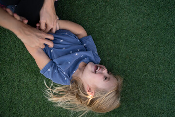 Niña rubia con ojos azules tumbada en el cesped riéndose porque unas manos le hacen cosquillas